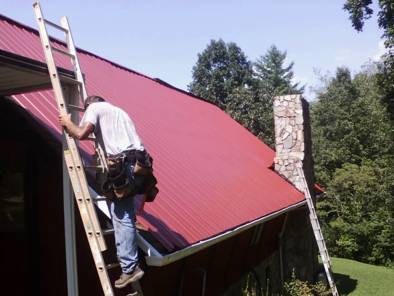 Roofer on a ladder beside a red metal roof and stone chimney — Soffit & Fascia Repair by Roofing Brothers in Rayland